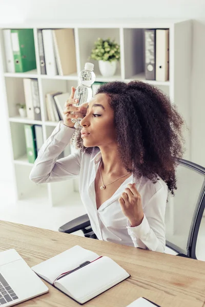 Exausto Africano americano empresária sofrendo de calor no escritório e segurando garrafa de plástico perto da cabeça — Fotografia de Stock