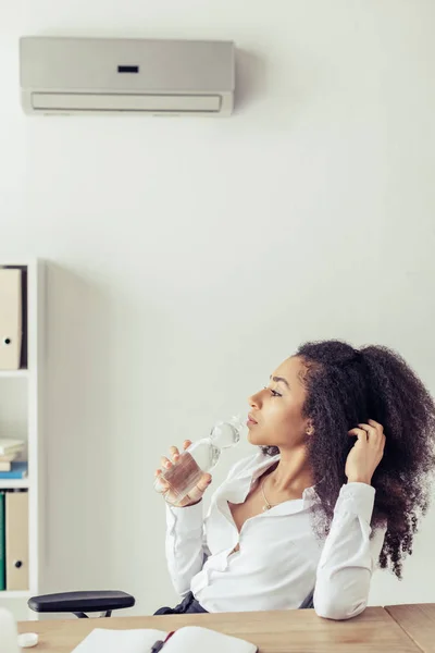 Pensativa mujer de negocios afroamericana beber agua mientras está sentado bajo el aire acondicionado en la oficina - foto de stock