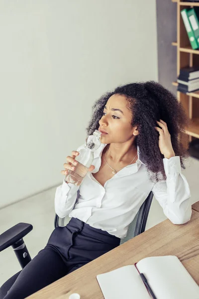 Pensierosa donna d'affari africana americana che beve acqua e distoglie lo sguardo mentre soffre di calore in ufficio — Foto stock