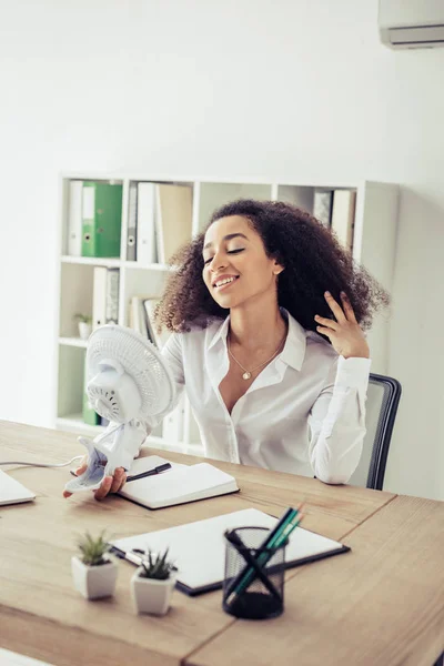 Cheerful african american businesswoman holding electric fun while suffering from heat in office — Stock Photo