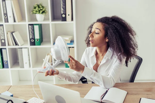 Mujer afroamericana joven sosteniendo ventilador de escritorio mientras está sentada en el lugar de trabajo y sufriendo de calor en la oficina - foto de stock