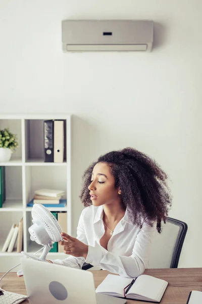 Afrikanisch-amerikanische Geschäftsfrau hält blasenden elektrischen Ventilator, während sie am Arbeitsplatz unter Klimaanlage sitzt — Stockfoto