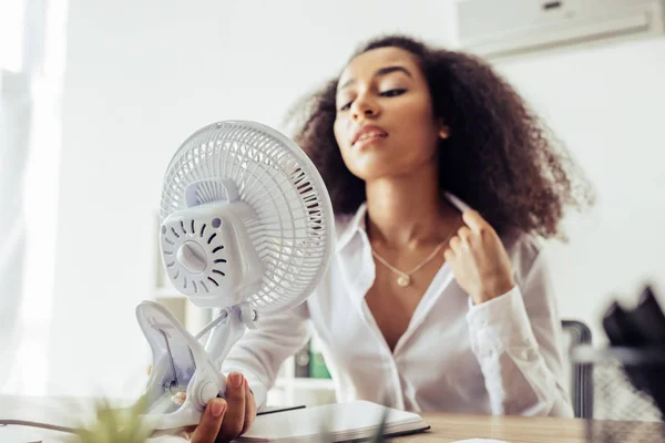 Focalizzazione selettiva di attraente donna d'affari africana americana con ventilatore elettrico mentre si siede sul posto di lavoro — Foto stock