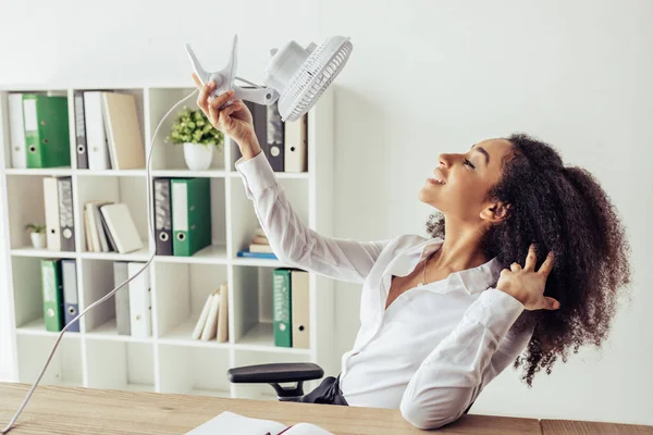 Cheerful african american businesswoman holding electric fan while suffering from heat in office — Stock Photo