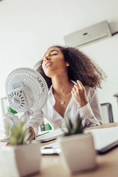 Selective focus of african american businesswoman holding electric fan while suffering from heat in office — Stock Photo