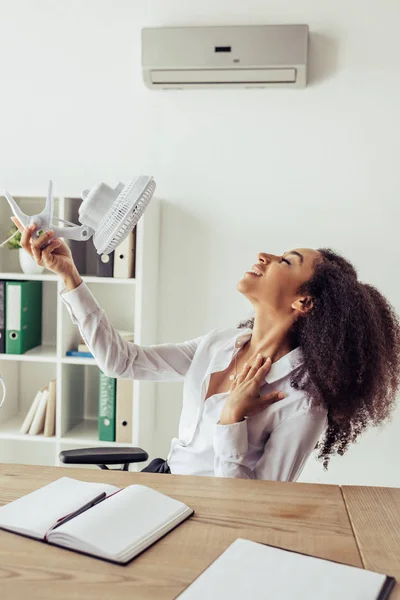 Pretty african american businesswoman sitting at workplace and holding electric fan while sitting at workplace in office — Stock Photo