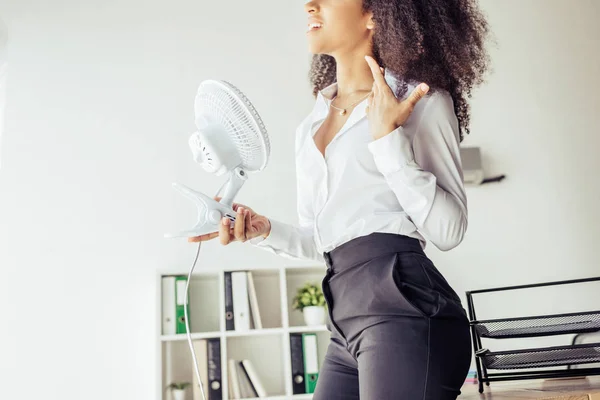 Vista parcial de la mujer de negocios afroamericana sosteniendo ventilador de escritorio mientras sufre de calor en la oficina - foto de stock