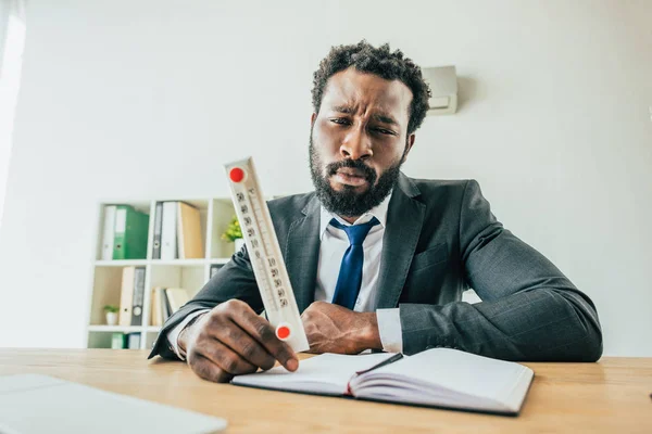 Infeliz empresário afro-americano segurando termômetro mostrando alta temperatura enquanto sentado no local de trabalho no escritório — Fotografia de Stock
