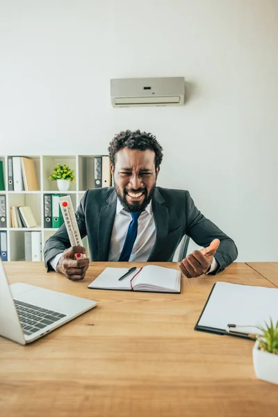 Irritated african american businessman holding thermometer and gesturing while suffering from summer heat in office — Stock Photo