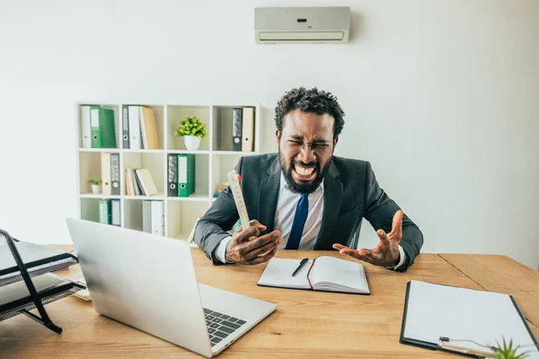 Hombre de negocios afroamericano enojado sosteniendo el termómetro y el gesto mientras está sentado en el lugar de trabajo en la oficina - foto de stock