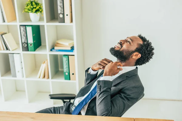 Hombre de negocios afroamericano agotado mirando hacia arriba y tocando la corbata mientras sufre de calor en la oficina - foto de stock