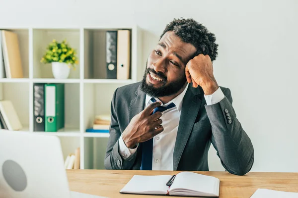 Unzufriedener afrikanisch-amerikanischer Geschäftsmann berührt Krawatte während er am Arbeitsplatz sitzt und unter Hitze im Büro leidet — Stockfoto