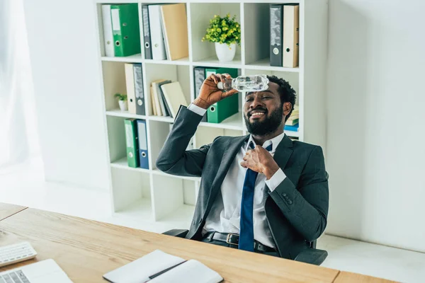 Hombre de negocios afroamericano sosteniendo botella de plástico con agua cerca de la cabeza mientras sufre de calor de verano en la oficina - foto de stock