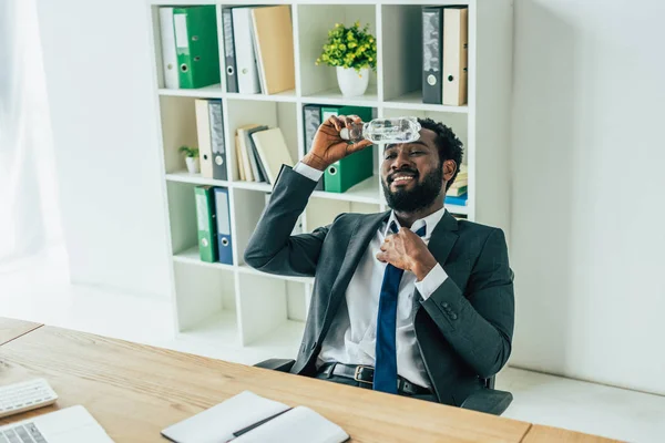 Homme d'affaires afro-américain tenant une bouteille en plastique avec de l'eau près de la tête tout en souffrant de chaleur estivale au bureau — Photo de stock