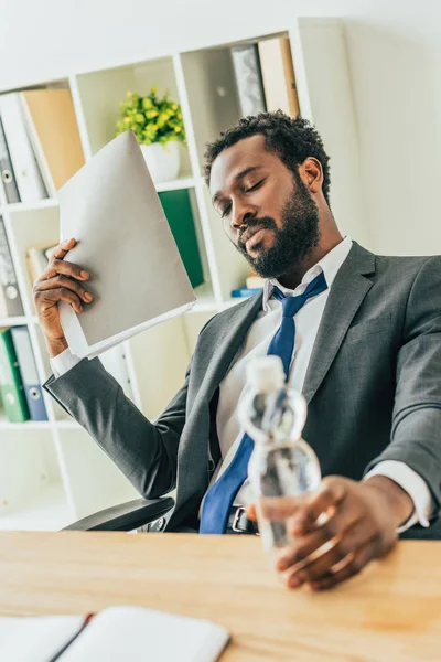 Exhausted african american businessman holding bottle of water and folder while suffering from heat in office — Stock Photo