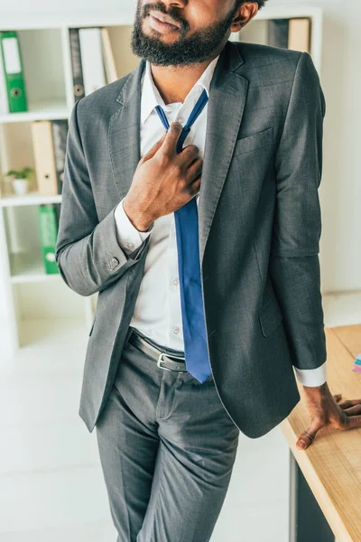 Abgeschnittene Ansicht eines afrikanisch-amerikanischen Geschäftsmannes, der am Arbeitsplatz steht und Krawatte berührt, während er im Büro unter Hitze leidet — Stockfoto