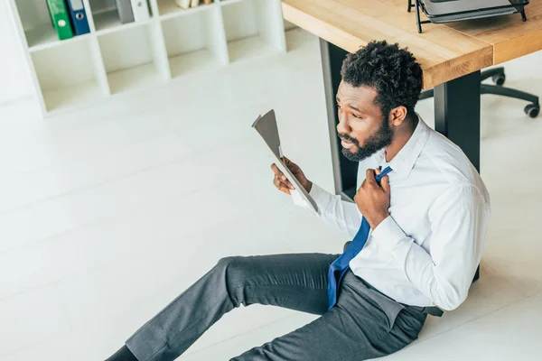 Joven hombre de negocios afroamericano sentado en el suelo y saludando con carpeta mientras sufre de calor en la oficina - foto de stock