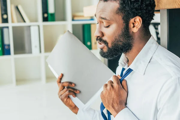 Dissatisfied african american businessman touching tie and waving with folder while suffering from heat in office — Stock Photo