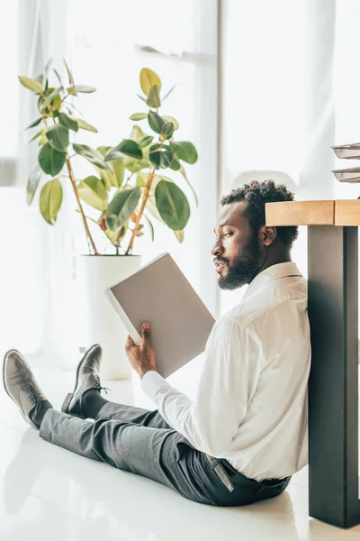 Hombre de negocios afroamericano insatisfecho sentado en el piso y saludando con carpeta mientras sufre de calor en la oficina - foto de stock