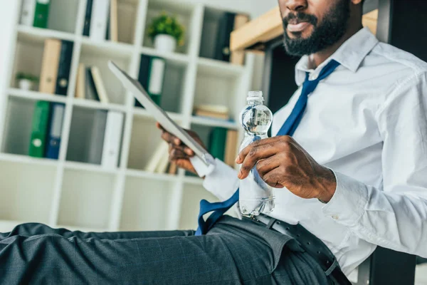 Cropped view of african american businessman holding bottle of water and waving with folder while sitting on floor in office — Stock Photo
