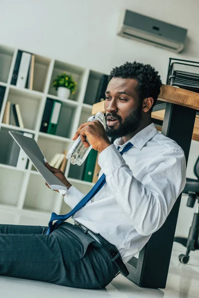 Young african american businessman sitting on floor and drinking water while suffering from heat in office — Stock Photo