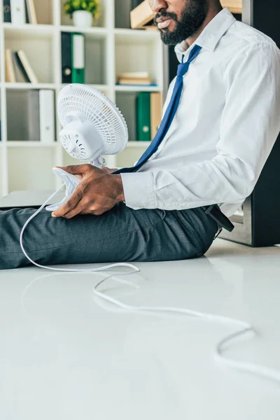 Vista parcial del empresario afroamericano sosteniendo ventilador eléctrico mientras está sentado en el piso en la oficina - foto de stock