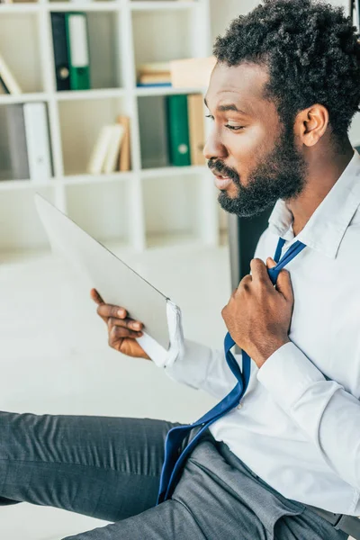 Hombre de negocios afroamericano agotado sentado en el suelo y saludando con carpeta mientras sufre de calor - foto de stock