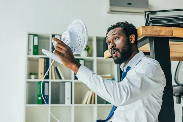 Young african american businessman holding blowing electric fan while sitting on floor and suffering from heat in office — Stock Photo