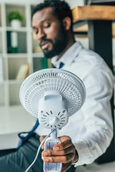 Foco seletivo do empresário afro-americano sentado no chão e segurando ventilador elétrico — Fotografia de Stock