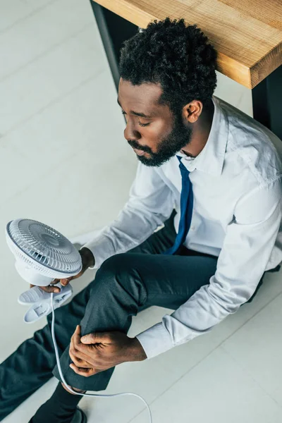 Blick auf einen afrikanisch-amerikanischen Geschäftsmann, der mit einem elektrischen Ventilator auf dem Boden sitzt und unter Hitze leidet — Stockfoto