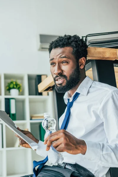 Unzufriedener afrikanisch-amerikanischer Geschäftsmann hält Flasche Wasser in der Hand, während er im Büro auf dem Boden sitzt — Stockfoto