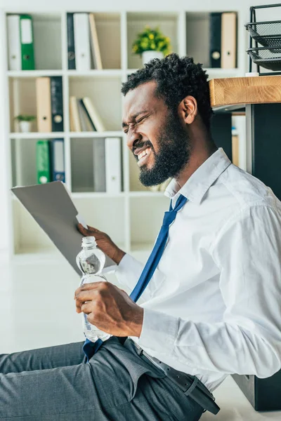 Hombre de negocios afroamericano agotado saludando con carpeta y sosteniendo la botella de agua mientras está sentado en el suelo y sufriendo de calor - foto de stock