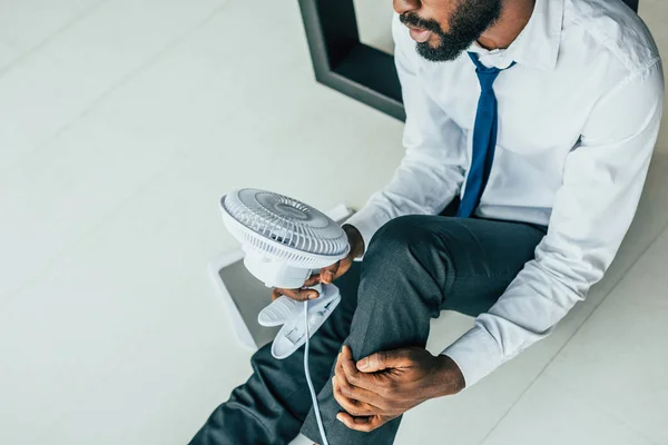 Abgeschnittene Ansicht eines afrikanisch-amerikanischen Geschäftsmannes, der auf dem Boden sitzt und einen blasenden elektrischen Ventilator hält — Stockfoto