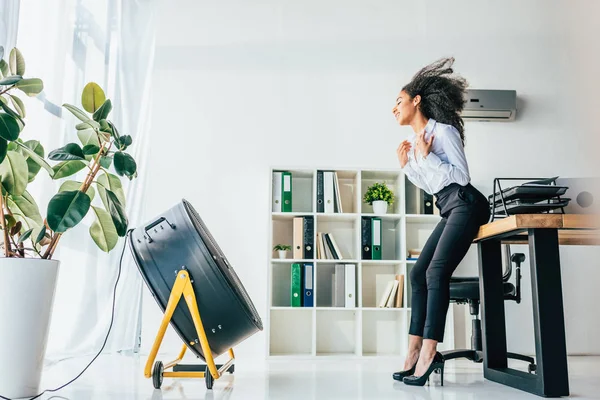 Sorridente donna d'affari afroamericana in piedi di fronte al ventilatore elettrico soffiante in ufficio — Foto stock