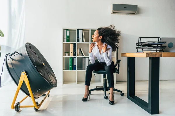 Pretty african american businesswoman sitting on office chair in front of blowing electric fan — Stock Photo