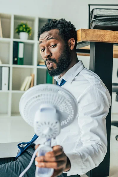 Schöner afrikanisch-amerikanischer Geschäftsmann mit blasendem Elektroventilator, während er im Büro auf dem Boden sitzt — Stockfoto