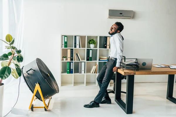 Exhausted african american businessman standing in front of electric ventilator in office — Stock Photo