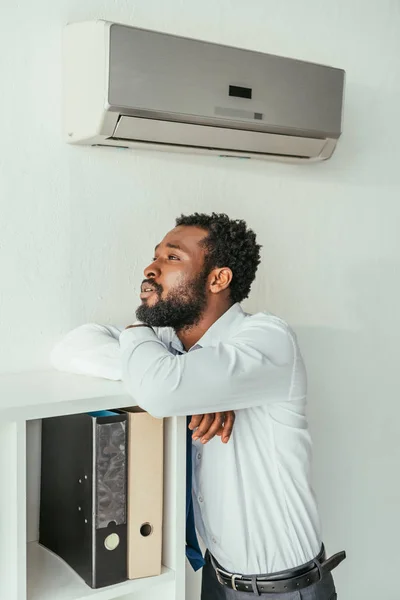 Pensive african american businessman standing under air conditioner and looking away in office — Stock Photo