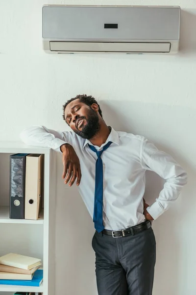 Hombre de negocios afroamericano agotado de pie bajo el acondicionador y cogido de la mano en la cadera - foto de stock