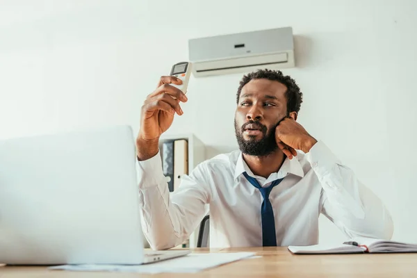 African american businessman holding air conditioner remote controller while suffering from heat in office — Stock Photo