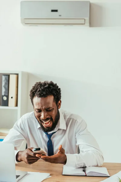 Irritado hombre de negocios afroamericano mirando el controlador remoto de aire acondicionado mientras sufre de calor en la oficina - foto de stock