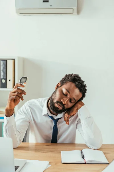 Hombre de negocios afroamericano agotado sentado con los ojos cerrados y sosteniendo el controlador remoto del aire acondicionado - foto de stock