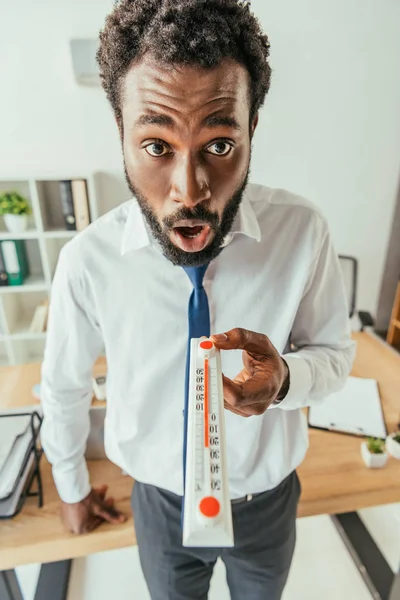 Шокований афроамериканський бізнесмен дивиться на камеру і показує термометр — стокове фото