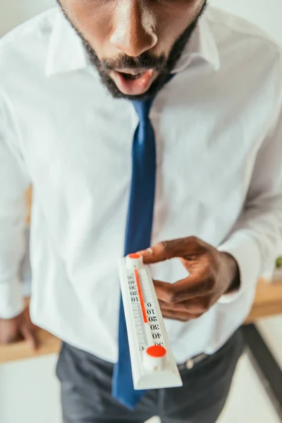 Cropped view of shocked african american businessman holding thermometer showing high temperature in office — Stock Photo