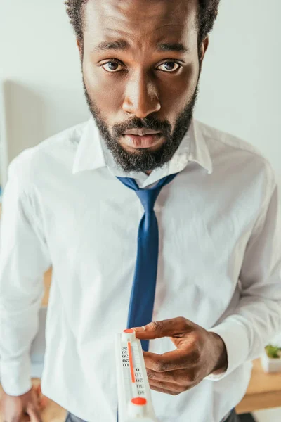 Виснажений афроамериканський бізнесмен тримає термометр і дивиться на камеру — стокове фото