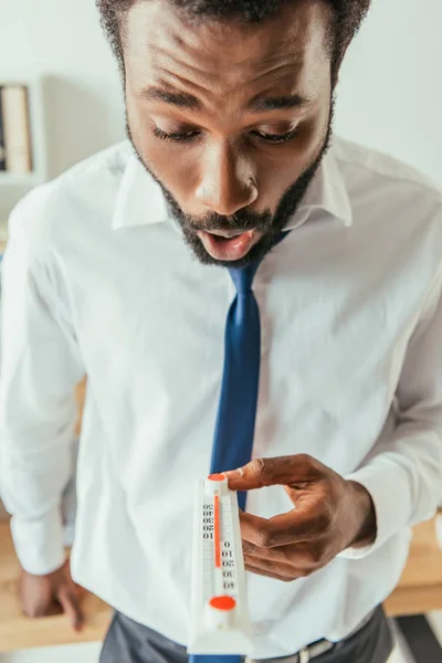 Шокированный африканский американский бизнесмен смотрит на термометр, показывающий высокую температуру в офисе — стоковое фото