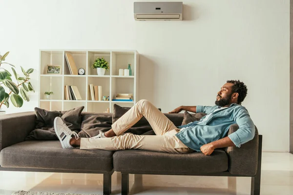 Молодой африканский американец лежит дома на диване, страдая от летней жары — стоковое фото