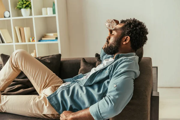 Hombre afroamericano exhausto acostado en el sofá con los ojos cerrados y sosteniendo botella con agua cerca de la cabeza mientras sufre de calor - foto de stock