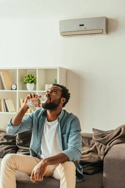 Hombre afroamericano que sufre de calor en casa y beber agua de botella de plástico - foto de stock