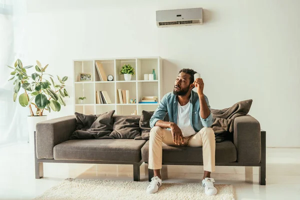 Tired african american man sitting on sofa and holding air conditioner remote controller while suffering from heat at home — Stock Photo